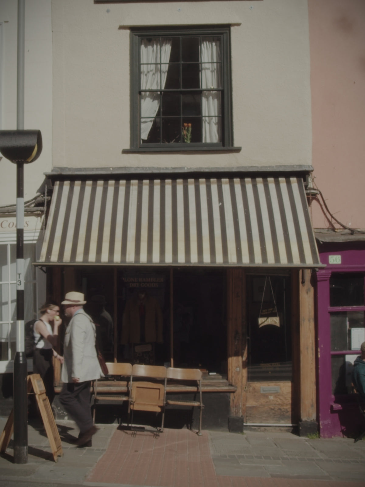 Shopfront with black and white striped awning, chairs and table outside, and two people walking past on the pavement.