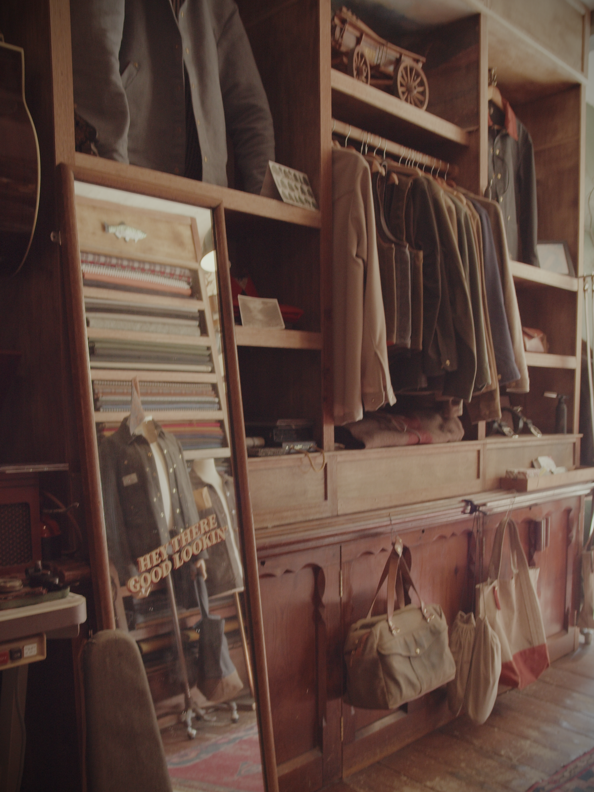 Inside the workshop with clothing rack, shelves, bags on display, and mirror reading “Hey there, good looking.”