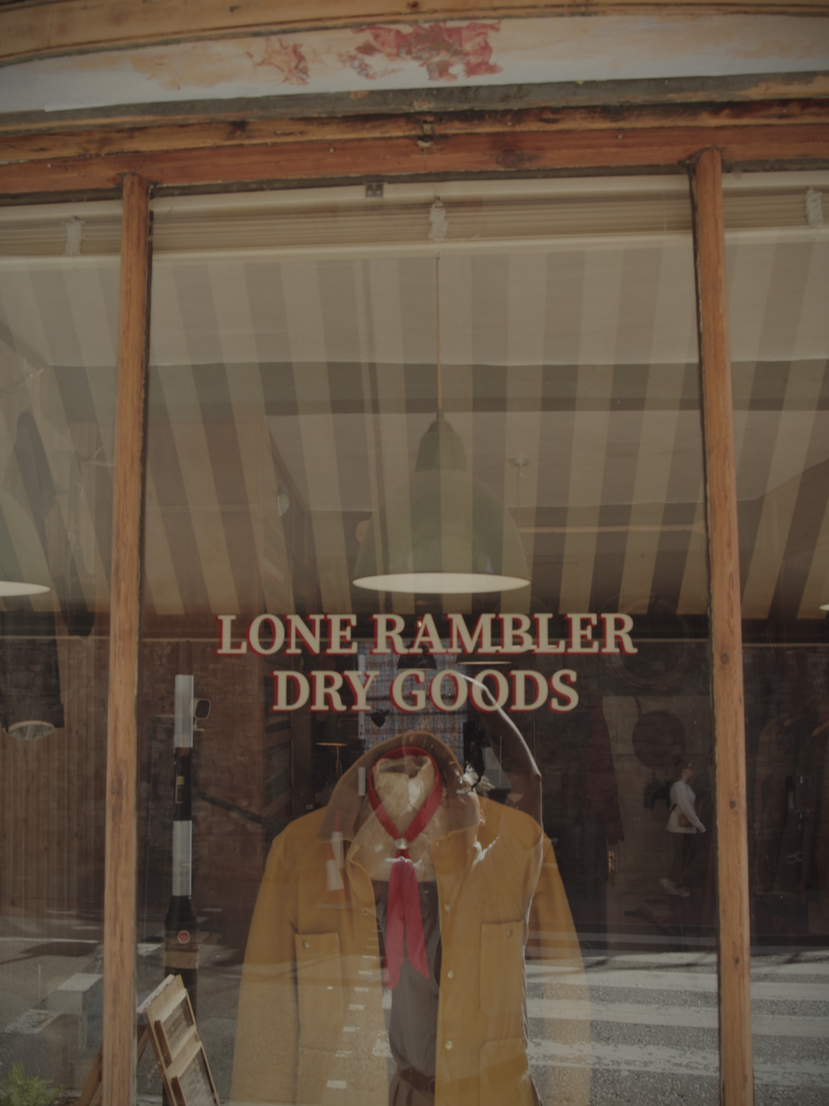 Shop window with “Lone Rambler Dry Goods” lettering in ebony and red, with interior visible and a painting of cowboys above the window.