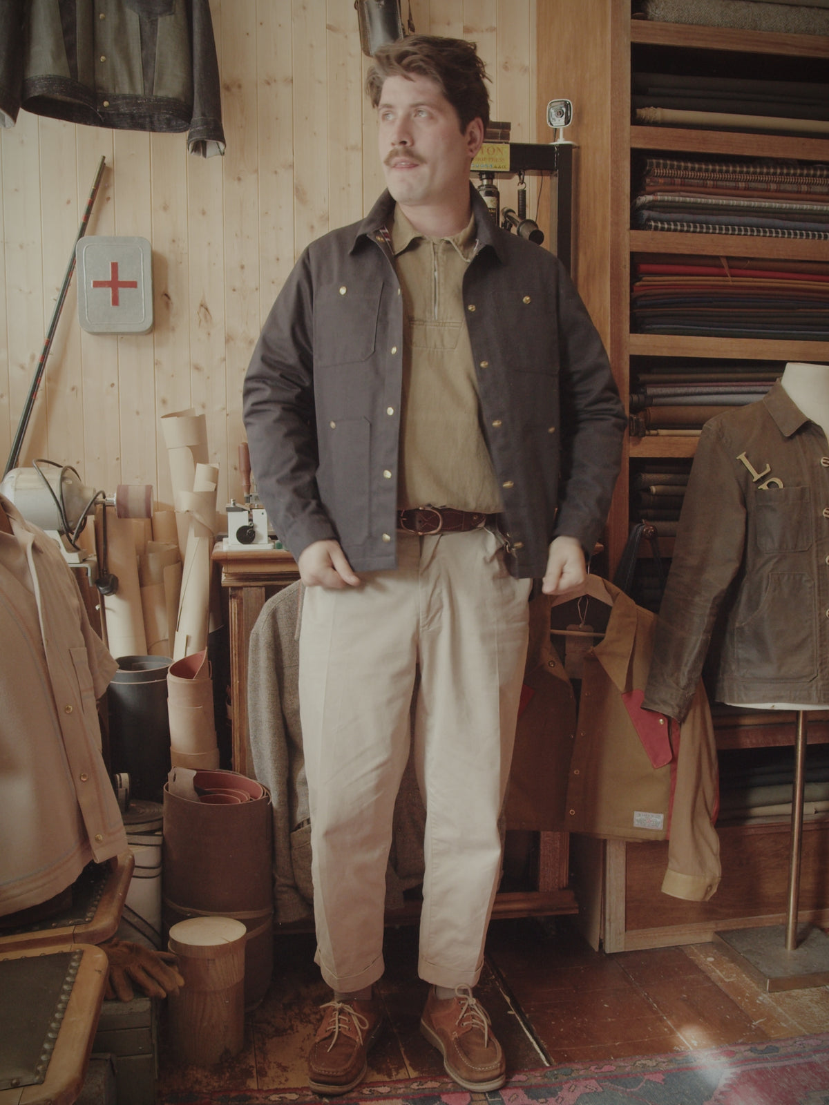 Man wearing Half-Moon jacket in dark waxed canvas inside workshop, full outfit view.